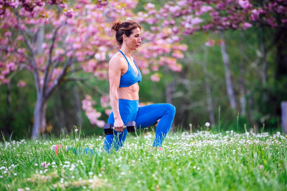 Woman kneeling and exercising with dumbbells in a grassy field with wildflowers and blooming pink trees outdoors, Calisthenics, USA.
