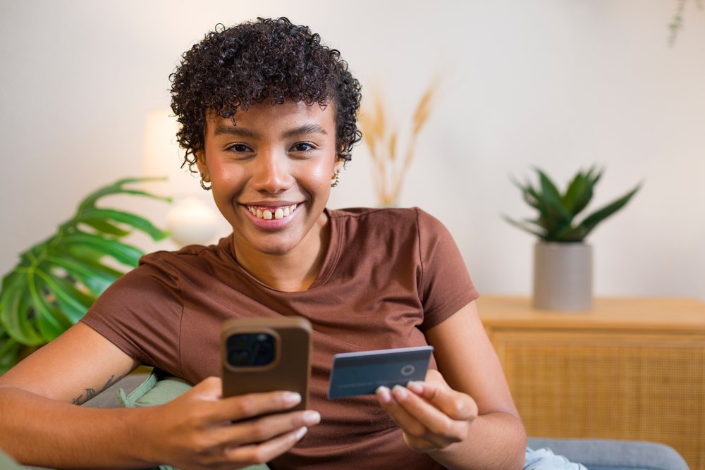 Young woman smiling and holding a smartphone and credit card in a modern home with potted plants nearby.