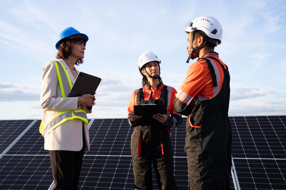 Three engineers in safety helmets discuss a solar energy project outdoors beside installed solar panels, Madrid, Spain.