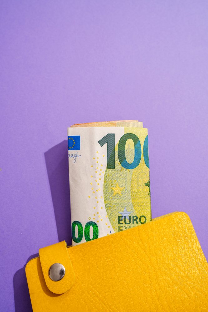 Rolled 100 euro banknote partially inserted in yellow wallet with metal snap against purple background, Studio Shot.