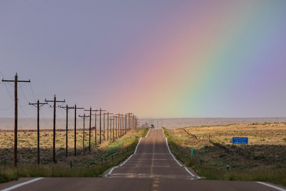 Vehicle drives on Highway 10, beneath a rainbow in a rural countryside setting with power lines and a weather sign nearby, Storm Chasing, Environment, Rocky Ford, Colorado, USA.