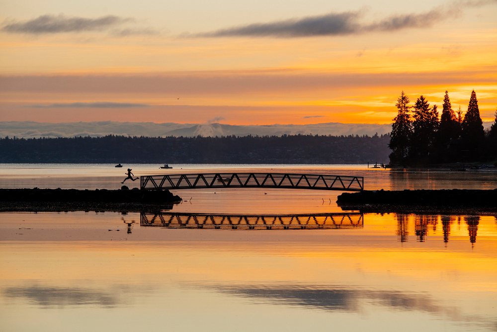 Silhouetted person jumping over rocks near a metal bridge on Bainbridge Island at sunset with calm water and tree reflections, WA, USA.