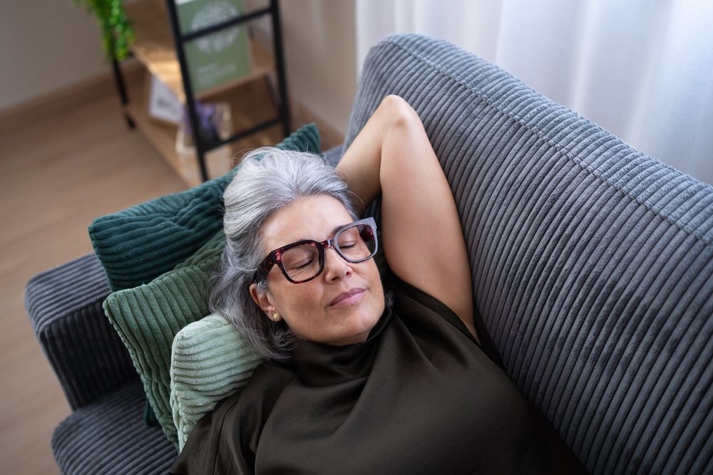 Middle-aged woman with gray hair wearing glasses rests with eyes closed on a gray sofa with green cushions indoors, Spain.
