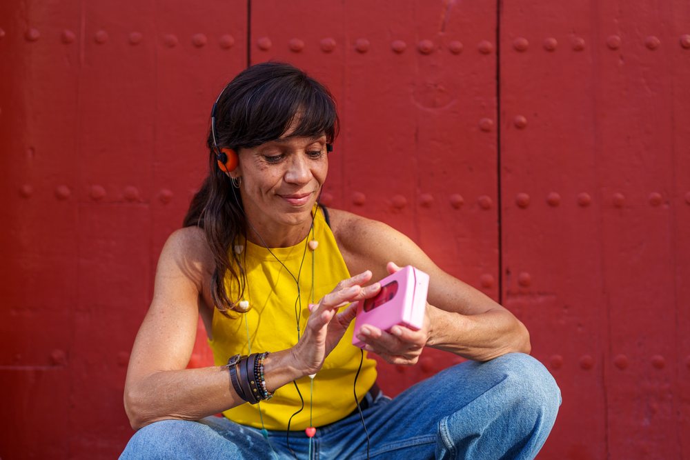 Woman sitting cross-legged outdoors against a red wooden door, wearing headphones and smiling while holding a music player, retro, vintage, Málaga, Spain.