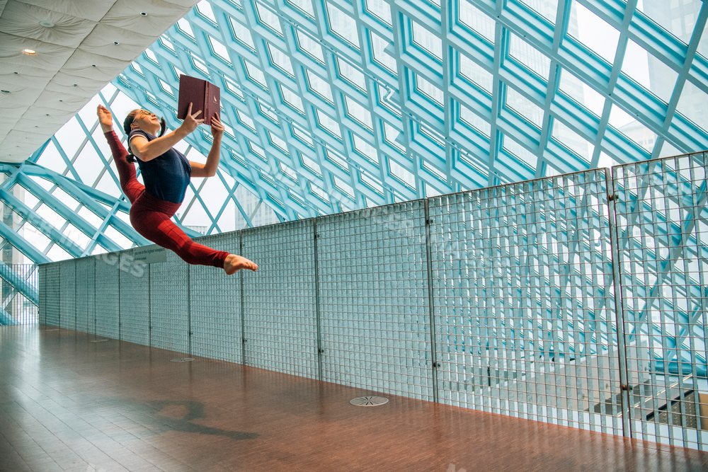 A woman jumps in a split leap holding a book in a library