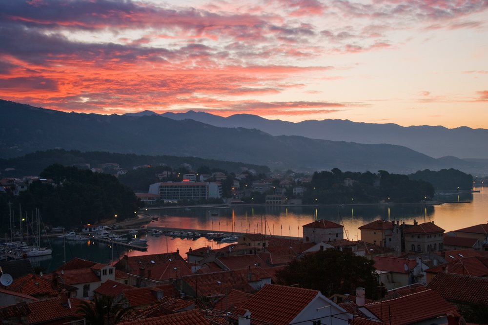 Town of Rab harbor at dawn showing boats in calm water with mountains and a red sunrise sky, Rab Island, Croatia.