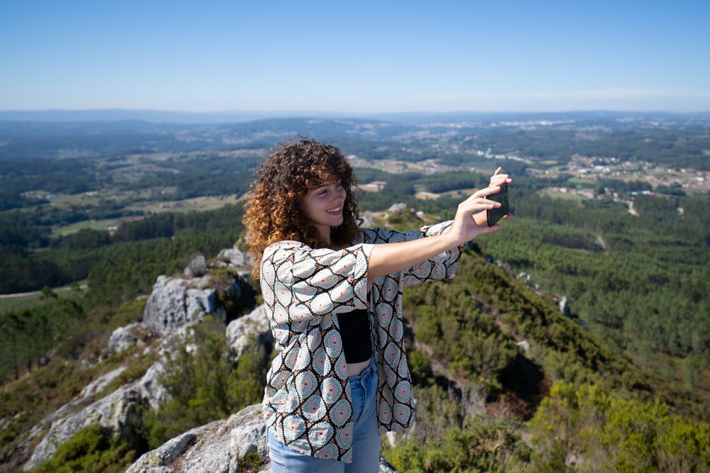 Woman taking a selfie on a rocky mountain peak overlooking a forest-covered valley, Pico Sacro, Galicia, Spain.