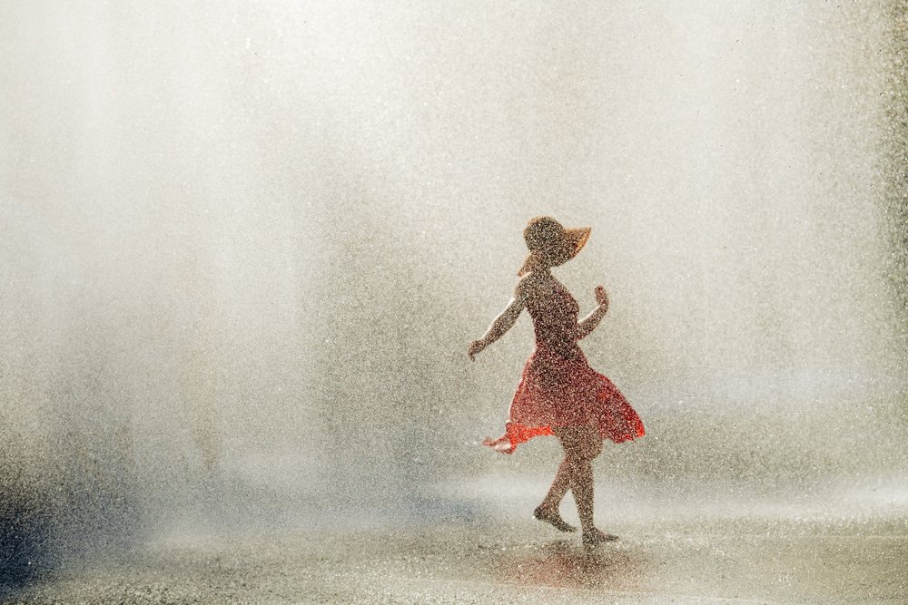 Woman wearing a red dress and sun hat dances barefoot outdoors in a summer water spray, USA.