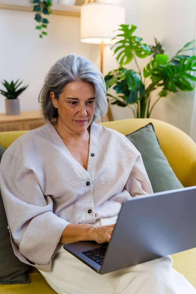 Mature woman with grey hair sits on a yellow couch using a laptop in a modern home interior, Spain.