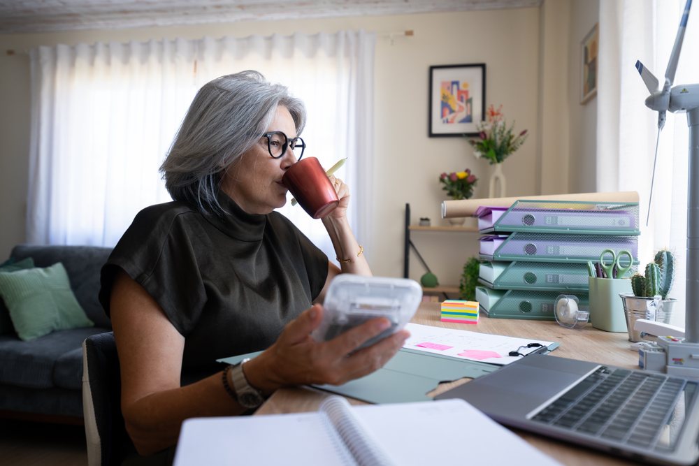 Woman with gray hair drinks coffee while working at a desk with laptop and calculator in a cozy home office, Spain.