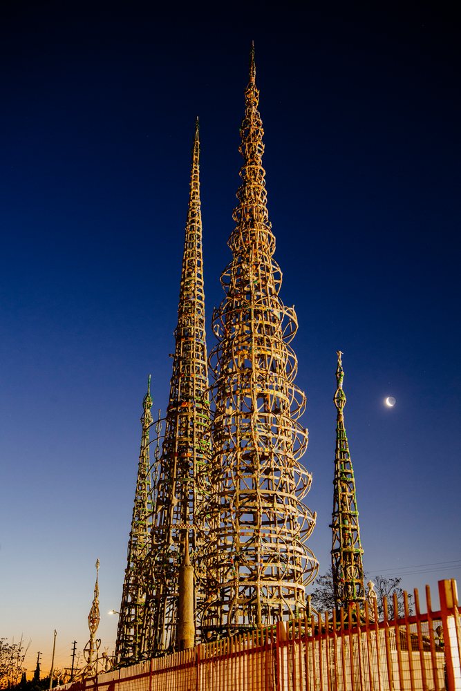 Watts Towers steel sculptures illuminated at twilight with a crescent moon and a fence in the foreground, California, USA.