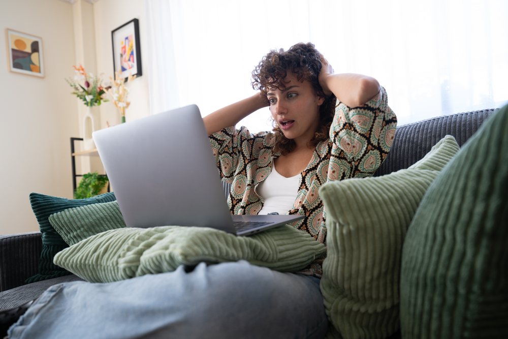 Woman sitting on a ribbed fabric couch using a laptop supported by pillows, with a surprised expression in a bright living room, home, girl, Spain.