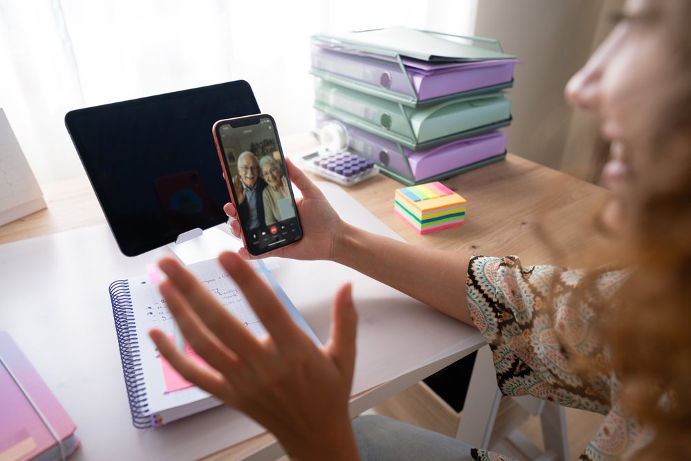 Woman at home in Spain holding smartphone and video calling an elderly couple at a desk with notebook and tablet.
