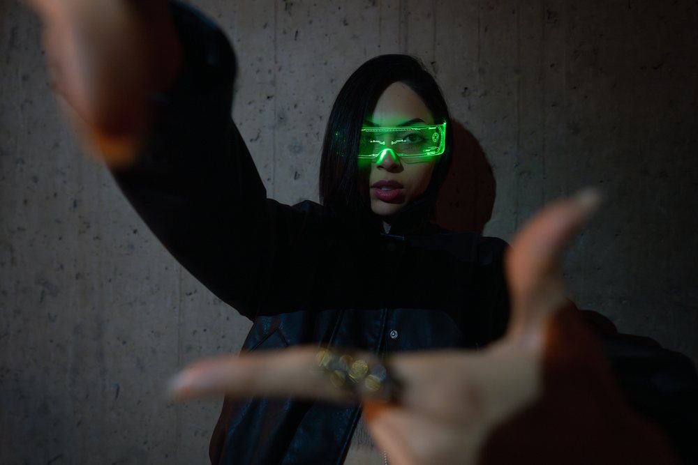 Woman wearing futuristic green glowing glasses and a black jacket poses indoors against a dark textured concrete wall, Madrid, Spain.