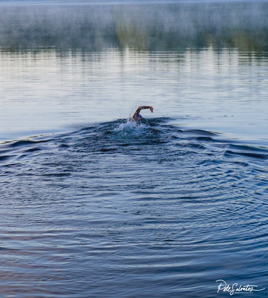 Swimmer performing front crawl stroke in calm lake water with morning mist, USA.