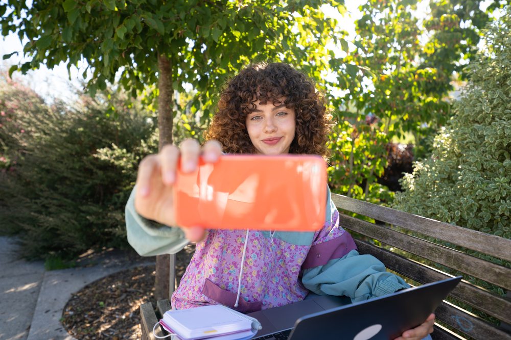 Woman with curly hair takes a selfie on a park bench outdoors with a laptop and notebook under a tree, Spain.