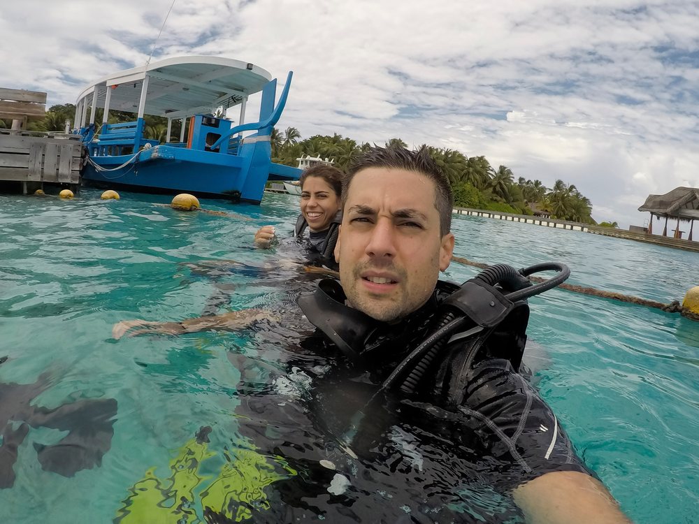 A man and woman wearing scuba diving gear in clear tropical waters near an island resort with a blue boat and palm trees in the background, Maldives.