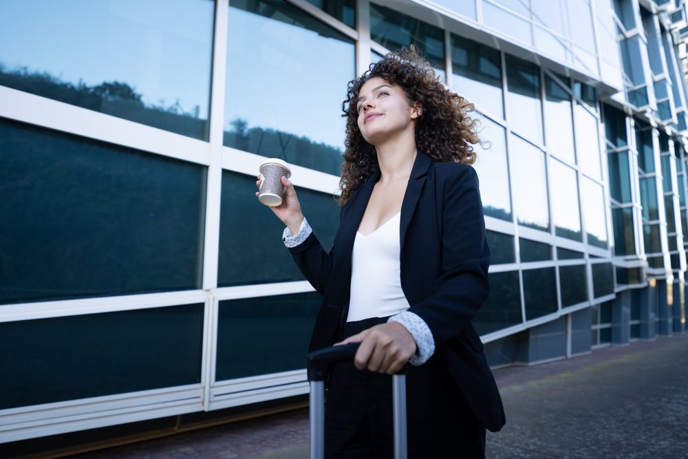 Businesswoman walking outdoors with rolling suitcase and coffee cup by office building with glass facade, business trip, Spain, Santiago De Compostela.