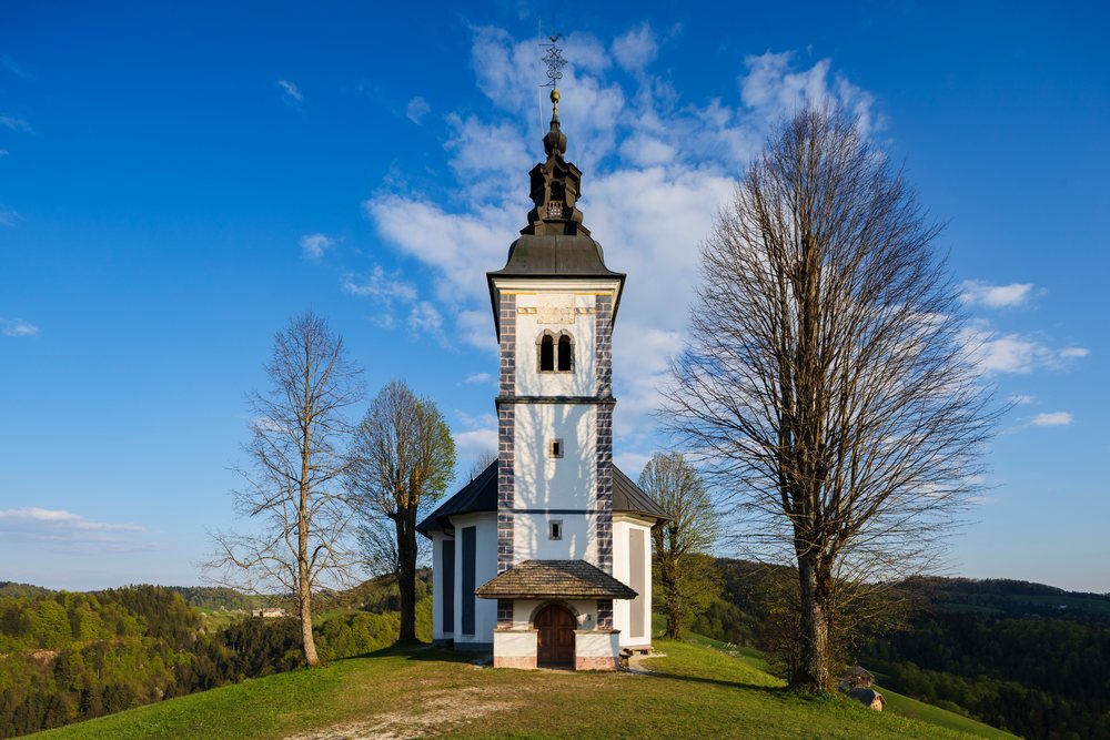 St Thomas's Church with a spired tower and Linden Trees on a grassy hilltop, Slovenia under a blue sky with white clouds, landscape, cityscape, Skofje Loka, Slovevia.