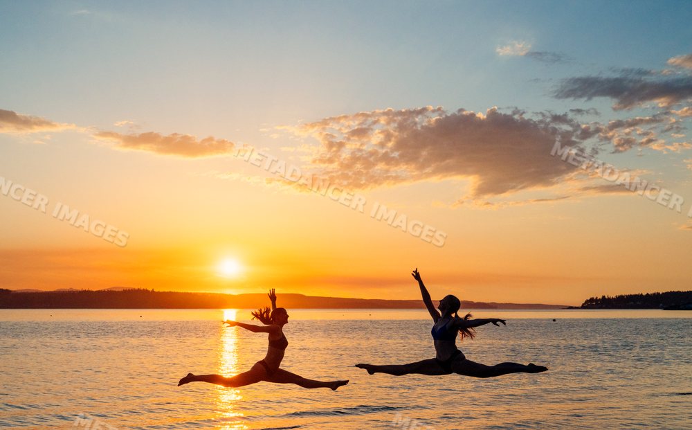 Two white women performing split leaps over water at sunset in WA, USA