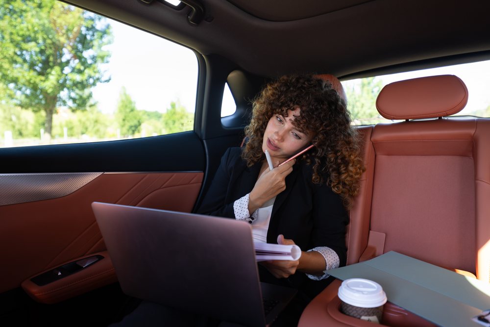 Businesswoman with curly hair working on a laptop and phone while holding a notebook in a car backseat, remote work, Spain.