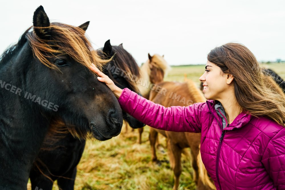 Young white woman happily pets black horse in outdoor pasture