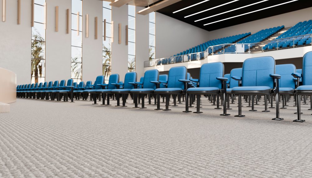 Rows of blue chairs with armrests face a stage inside a modern, spacious conference hall.