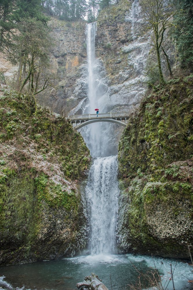Waterfall cascades behind a stone bridge with a person holding an umbrella, Multnomah Falls, Bridal Veil, Oregon.