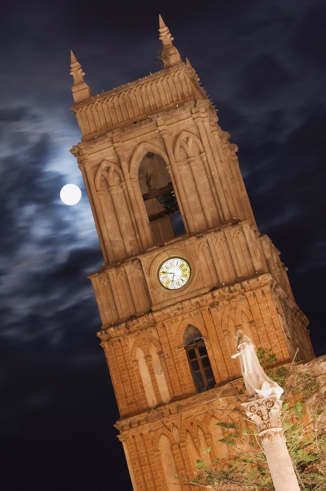 Illuminated clock tower with a statue on a stone column under a moonlit cloudy night sky, San Miguel de Allende, Mexico.