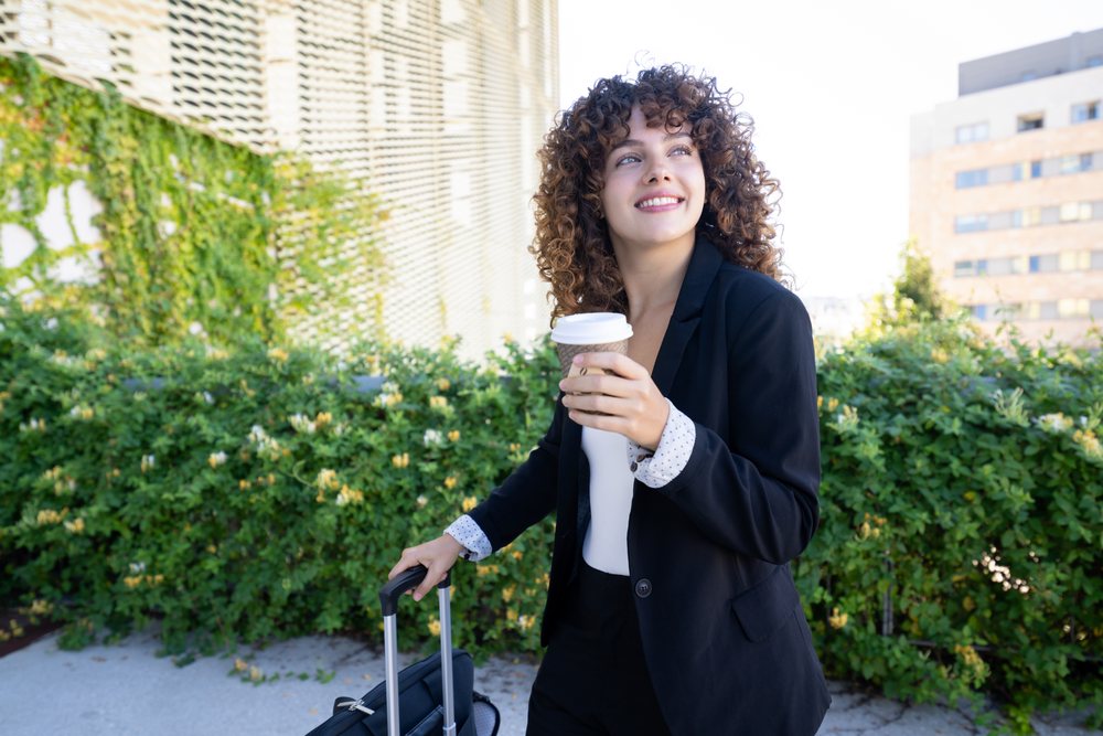 Woman wearing business attire holds a paper coffee cup and rolling suitcase outdoors in an urban environment with greenery and buildings visible, Spain.