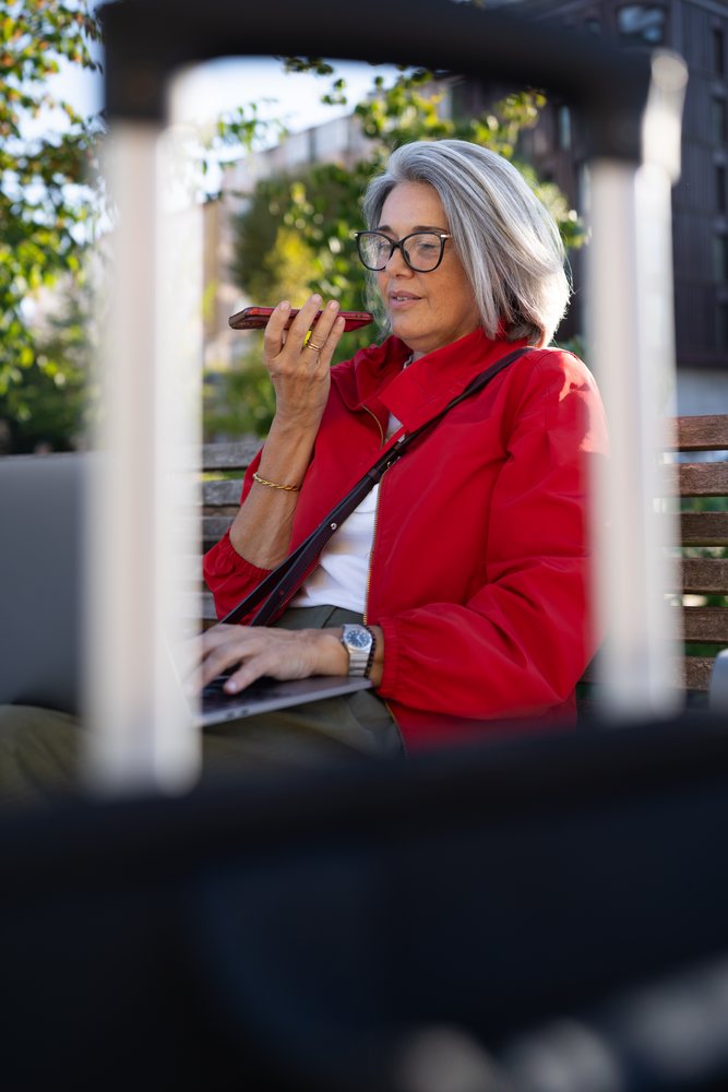 Woman wearing a red jacket talks on a smartphone while using a laptop on a bench outdoors in a park, Spain.