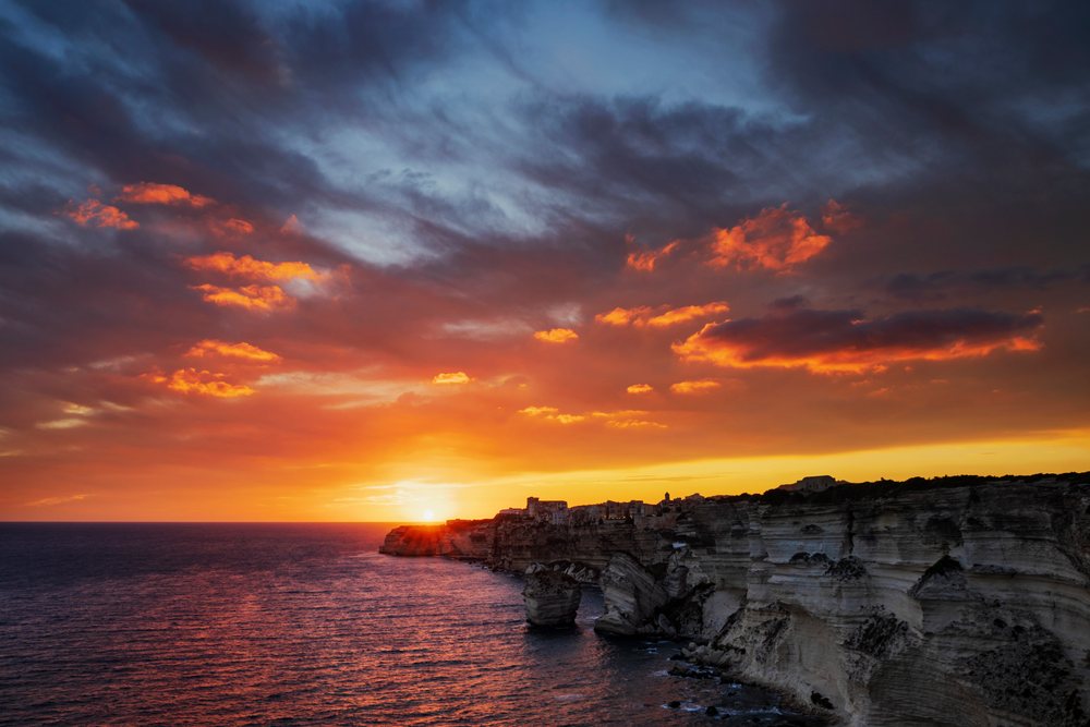 Sunset over rocky coastal cliffs and calm sea, with dramatic clouds and buildings atop the cliffs, Travel Photography, Bonifacio, Corsica, France.