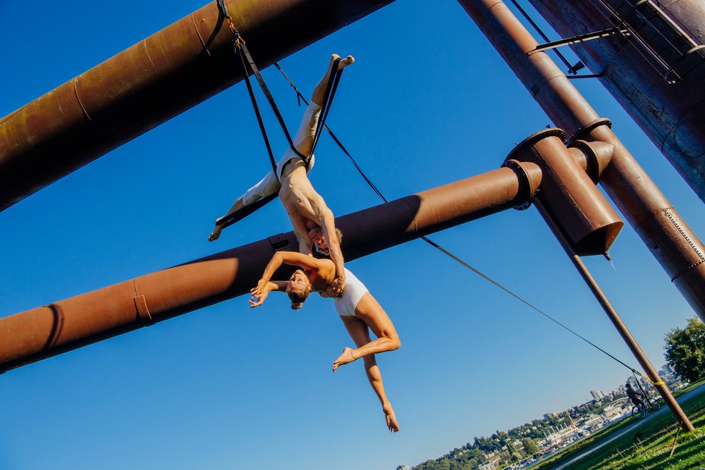 Two acrobats in white leotards perform an aerial routine on suspension straps against rusted industrial pipes in a Seattle urban park, WA, USA.