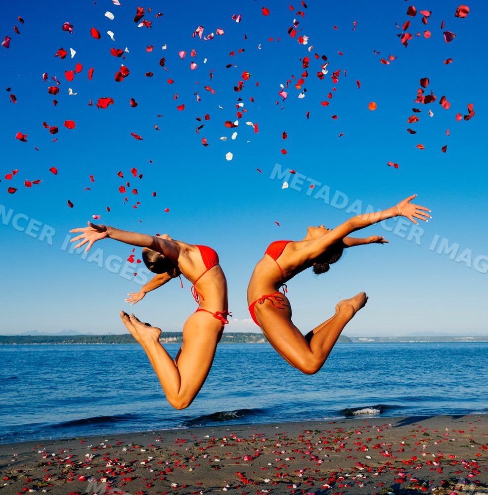 Two women in red swimwear jump on a beach with rose petals in wa USA