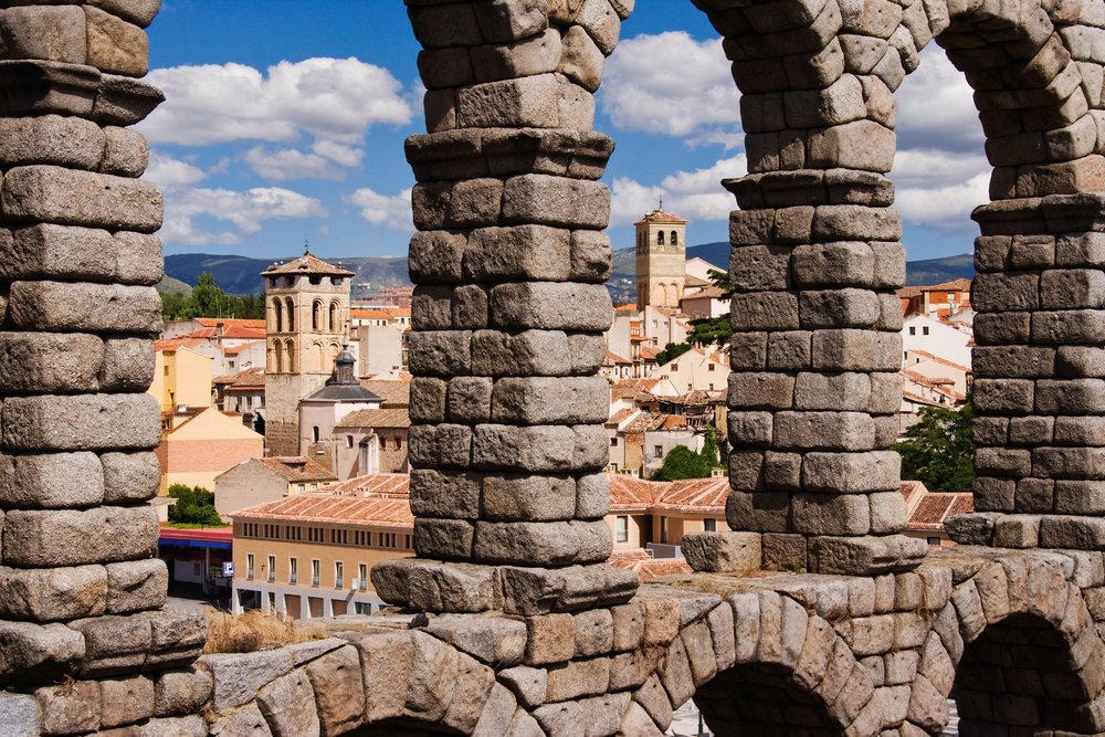 Roman aqueduct stone arches frame Segovia's historic cityscape beneath a blue sky with clouds and mountains, Spain.