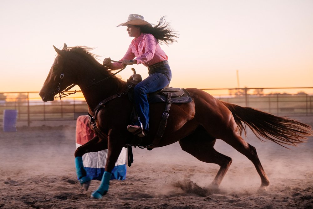 Cowgirl wearing a hat riding a galloping horse past barrels in a dusty outdoor arena at sunset, Southwest USA.