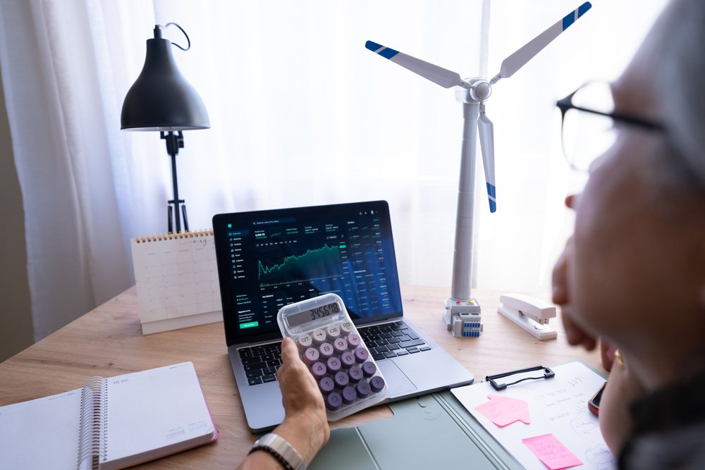 Person holding a calculator while viewing financial charts on a laptop at a desk with a wind turbine model and office supplies, Spain.