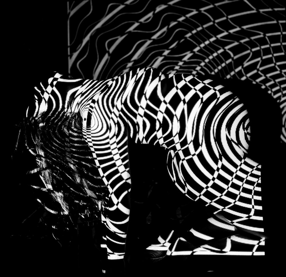 Woman kneels with an abstract black and white striped pattern projected onto her body against a dark background, pregnancy.