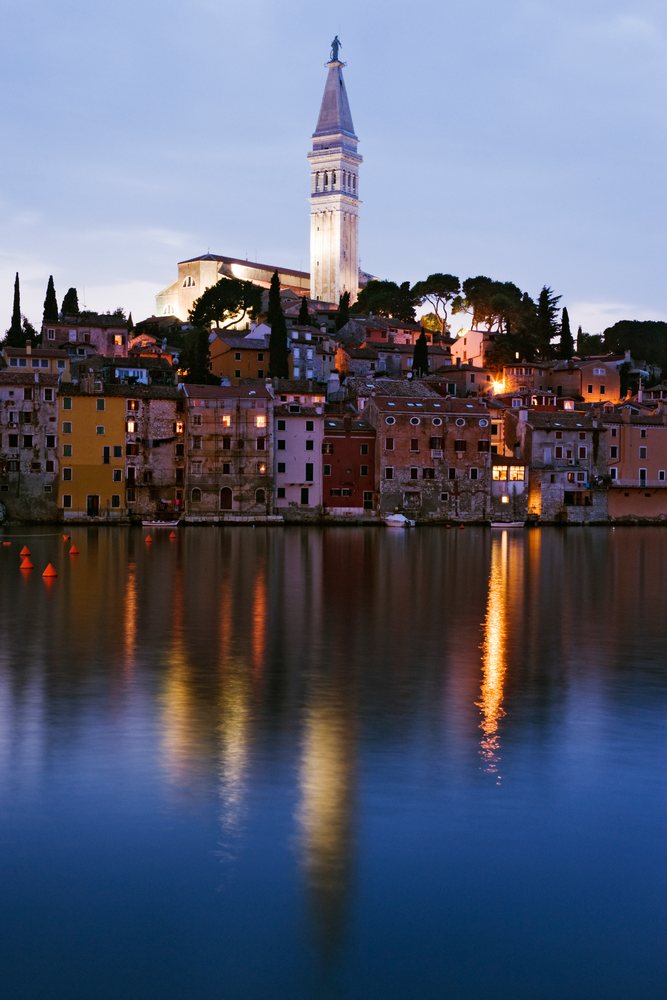 The town of Rovinj waterfront with illuminated buildings and church tower reflected on calm water, Sunset, Croatia.