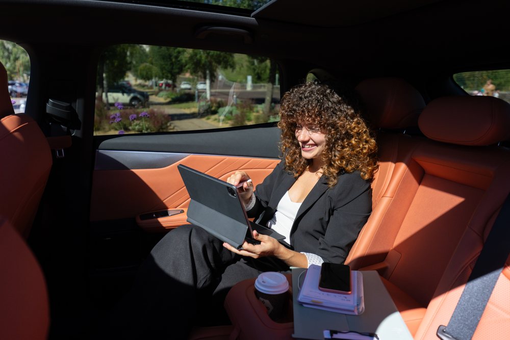 Businesswoman with curly hair works on a tablet inside a car's back seat near an urban park, with a coffee cup and notebook visible, remote work, Spain.