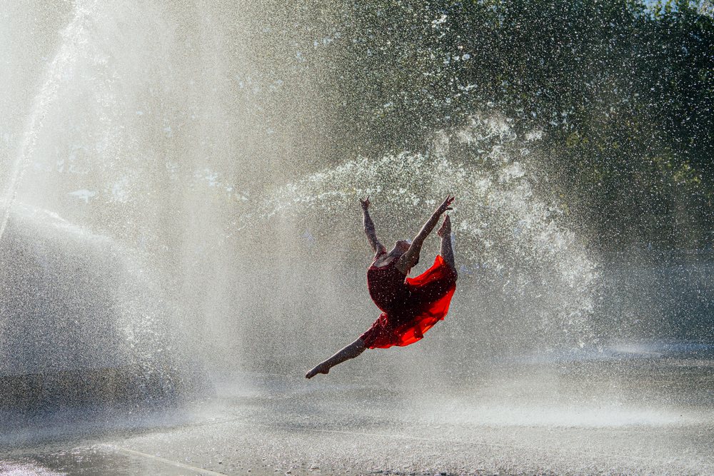 Dancer in a red dress leaping through water spray from an outdoor fountain on a sunny summer day, USA.
