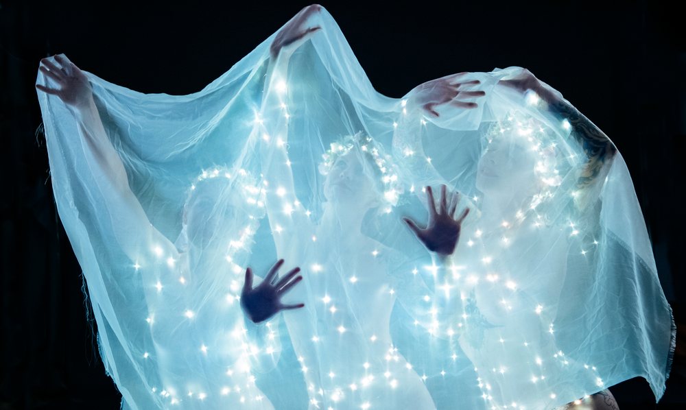 Three human figures draped in illuminated veils with fairy lights and head wreaths pose with spread hands against a dark background in an artistic performance, USA.