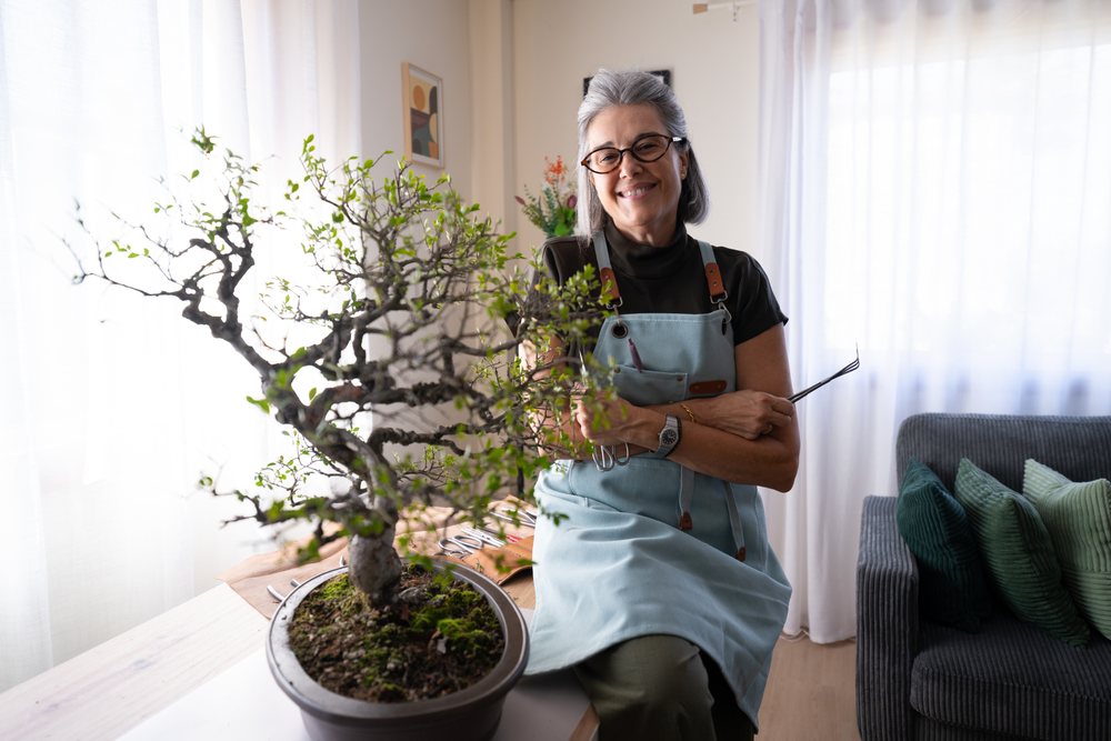 Woman with glasses and gray hair smiles while caring for a bonsai tree indoors in a living room, Spain.