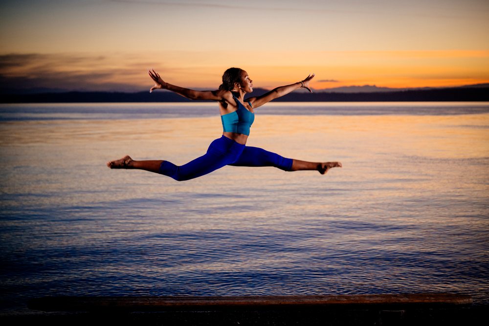 Woman performing a jumping split leap in athletic attire at sunset by a lake, yoga, acrobatics, USA.