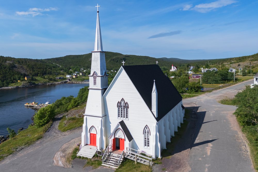 St Peter & Paul Roman Catholic Church with a white exterior, black roof, and red doors beside a road near a harbor, Landscape, Cityscape, Lifestyle, King's Cove, Newfoundland & Labrador, Canada.