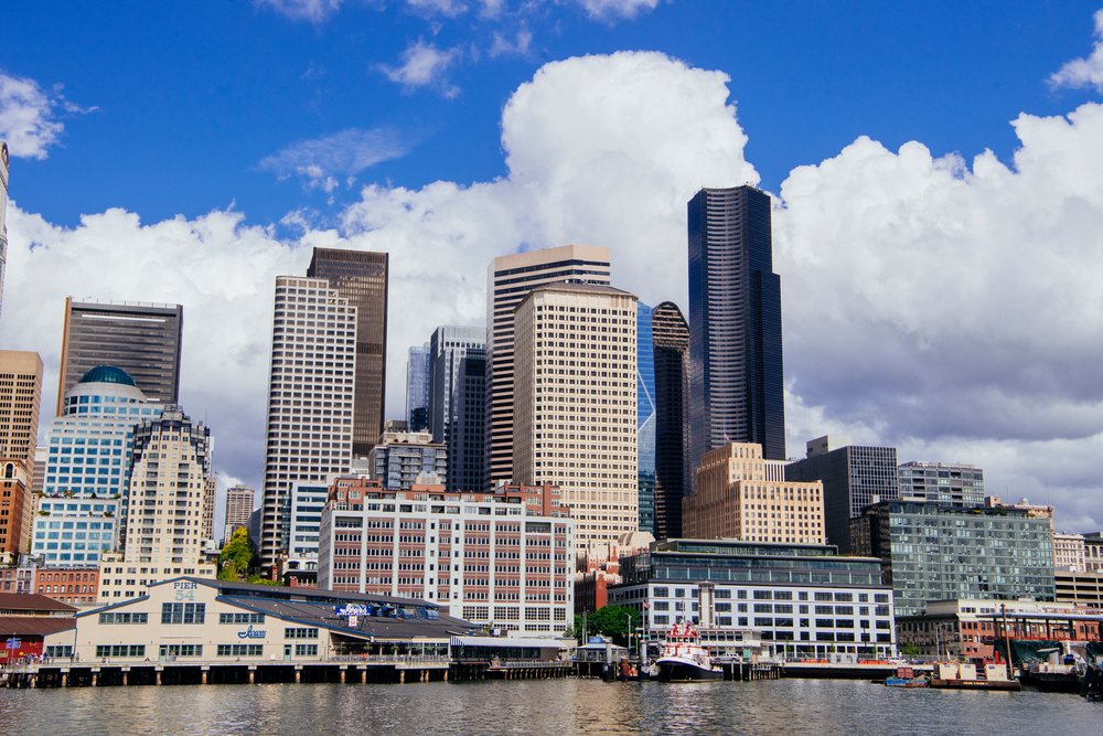 Seattle skyline featuring waterfront, Pier 54 buildings, boats, and reflections under a partly cloudy blue sky, USA.