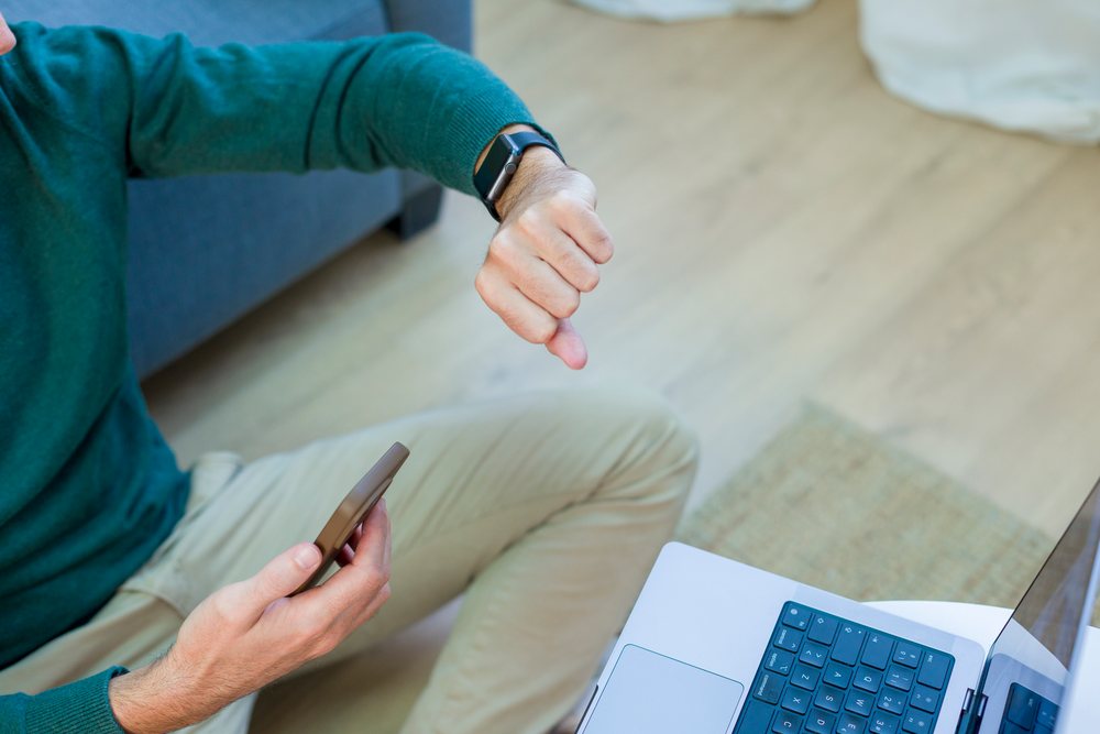 Man wearing a smartwatch and holding a smartphone while sitting indoors near a laptop at home using digital devices, Technology, young adult.