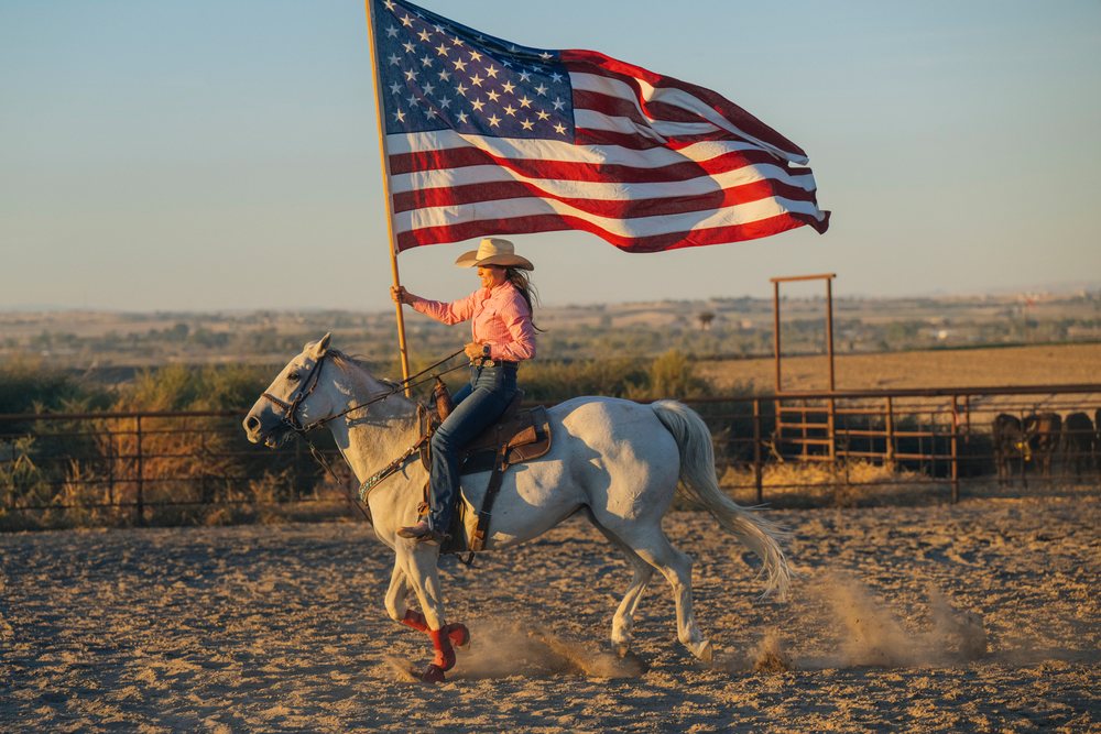 Woman wearing cowboy hat rides a saddled white horse holding an American flag in a fenced ranch corral at sunset, Southwest USA.