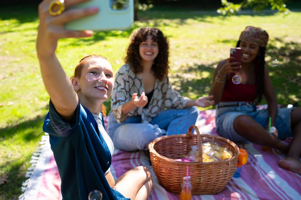 Three friends sit on a picnic blanket in a grassy park, enjoying drinks, snacks, and taking selfies on a sunny day, Spain.