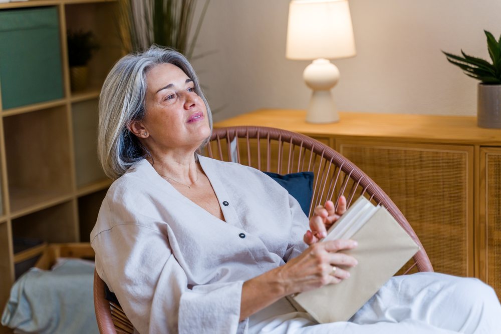 Woman with gray hair reading a book while sitting in a modern chair indoors near a lamp and plant, Spain.